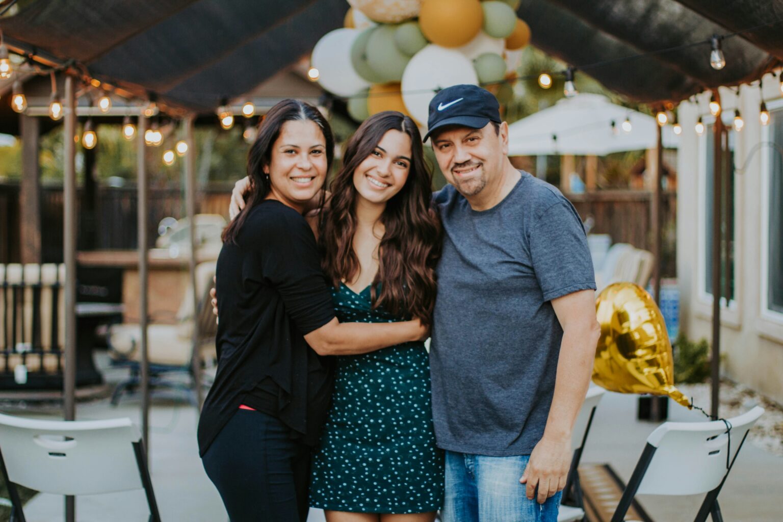 A family of three smiling together in a backyard decorated with balloons and string lights